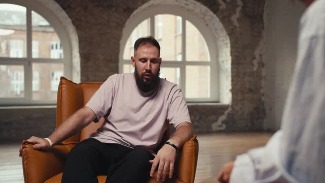 An excited man with a beard in a pink shirt talks about his problems and questions to a psychologist. A young man sits on a brown chair and communicates with a psychologist in a special room made of bricks