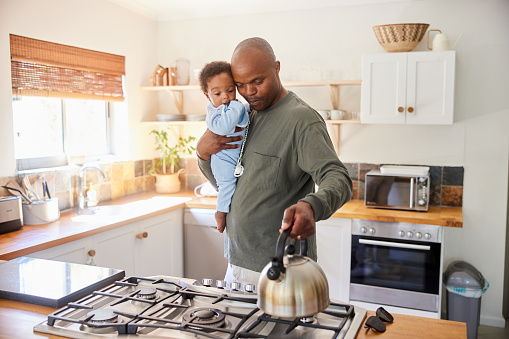 Smiling father boiling water in a kettle while standing with his baby boy in his arms in their kitchen at home