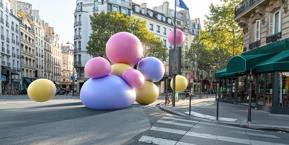 An abstract image of ten huge blue, pink and yellow balloons or spheres falling onto an empty city street in an older downtown high street location with shops and restaurants in the morning/afternoon on a sunny day.