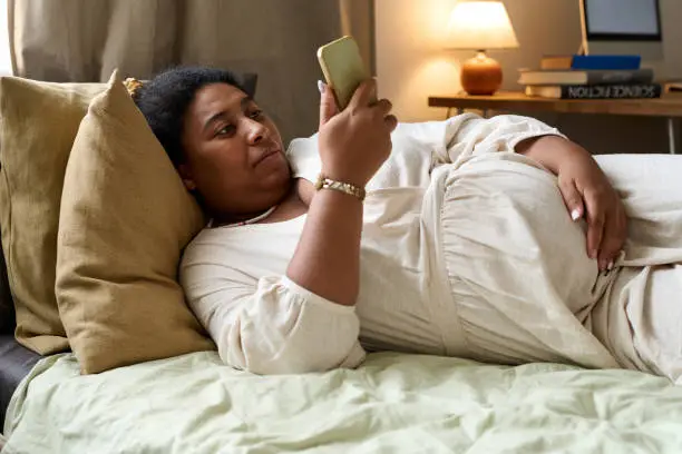 African American pregnant woman surfing the net on her mobile phone while lying on bed in the room