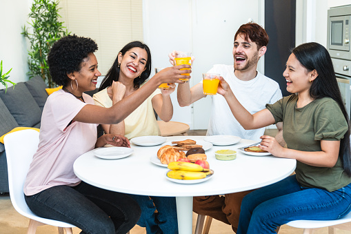 Diverse group of four laughing over breakfast, toasting with juice in a cozy kitchen setting.