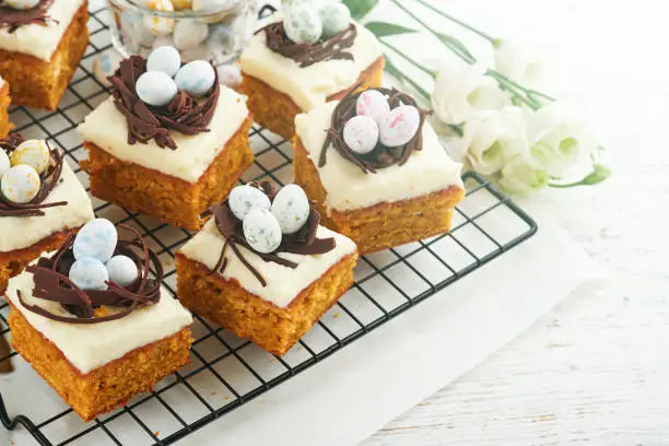 Photo of Easter carrot cake bars decorated with chocolate nest and chocolate candy eggs blossoming cherry or apple flowers on rustic light wooden backgrounds. Easter holiday meal. Traditional Easter food.