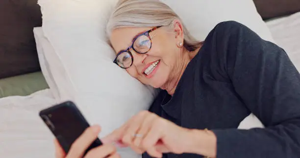 Photo of Happy, senior woman and reading with phone in bed or streaming funny, comedy or meme on social media. Elderly person, smile or relax with cellphone at night in bedroom with communication or chat