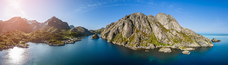 Panoramic drone view of Nusfjord in Lofoten, Norway, captures the vibrant blue waters and rugged cliffs under the summer sun, with a scattering of traditional Norwegian houses along the shoreline