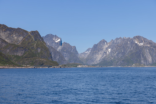 Norwegian panorama presents a tranquil North Sea tenderly embracing grand mountain ridge, with a charming harbor of Moskenes town perched along Lofoten Island's coast, all beneath a vast, azure sky on a serene summer day
