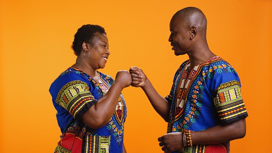 Young african american people greeting with fist bump on camera, saying hi to each other with modern gesture. Smiling husband and wife in traditional apparel posing in studio, cool salute.