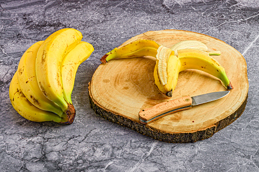 A ripe, yellow banana sits partially peeled on a wooden board, alongside a bunch of unpeeled bananas