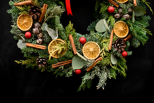 Cropped photo of Christmas wreath decorated with red ribbon, pine cones, red small balls, dried orange slices and vanilla sticks hanging on a hook on a black background