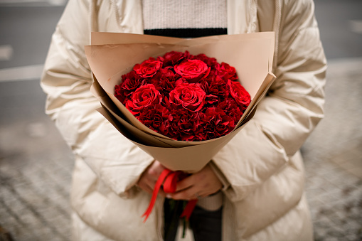 Large bouquet of red roses is held by female hands in winter clothes on a blurred street background