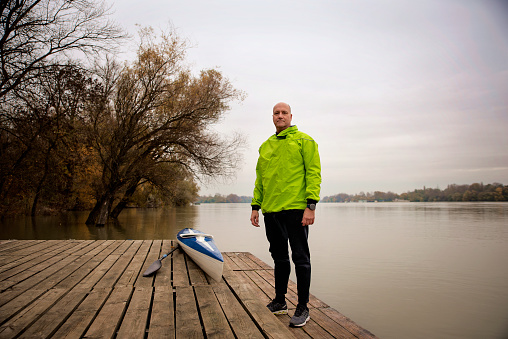 A middle-aged man standing on the jetty and ready to go kayaking. Active sporty man wearing dry suit and looking at camera.