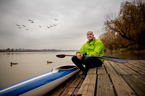A middle-aged man sitting on the jetty and ready to go kayaking. Active sporty man wearing dry suit and looking away.