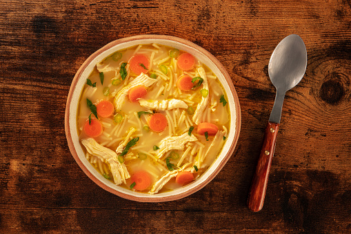 Chicken noodle soup with vegetables, a bowl of healthy stock with a spoon, top shot on a rustic wooden table, winter home cooking