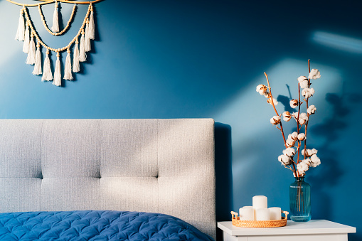Stylish modern bedroom in dark colors. Cozy interior with navy blue walls, home decor. Bed with gray fabric headboard, blue blanket, bedside table, vase with natural cotton flowers, candles on tray.