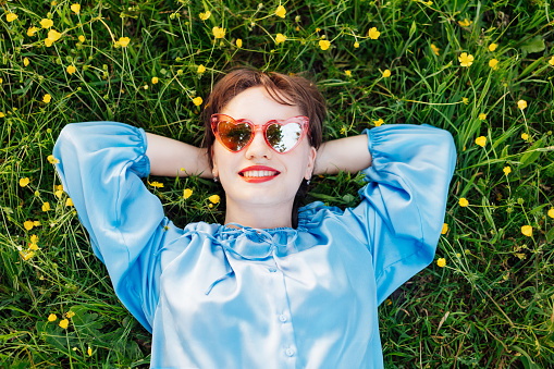 Top view happy young woman in blue blouse lays on a green grass lawn with yellow flowers, looks up and smiles. Business woman enjoy the moment, relaxing outdoors. Spring or summer season concept