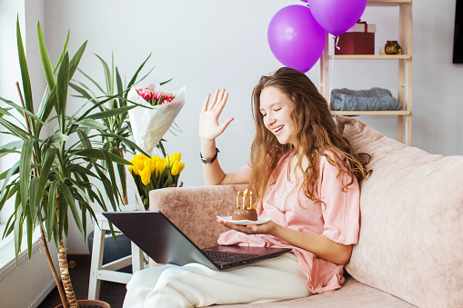 Happy young woman virtual meeting birthday party online together with her friend in video conference with laptop for online meeting in video call. Teen girl with birthday cake at home