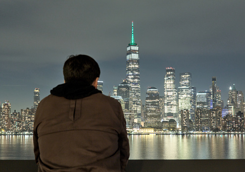 man looking at nyc skyline (from behind, silhouette) wearing hoodie and brown jacket (manhattan skyscrapers, world trade center, hudson river, waterfront view) travel destination new york city