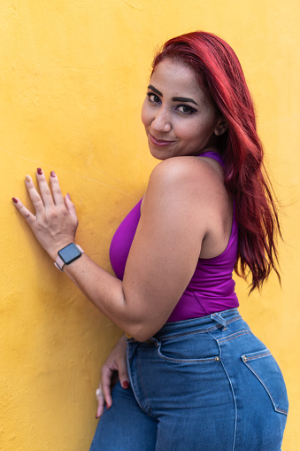 A red-haired woman in a sleeveless purple top leans playfully against a vibrant yellow wall.