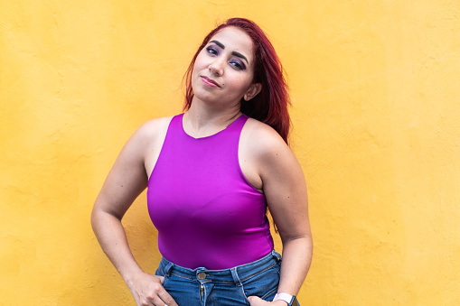 A woman with red hair in a vibrant fuchsia tank top, hands on hips, against a yellow backdrop.