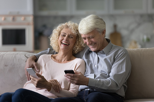 Cheerful senior married couple using applications, virtual services on smartphones for Internet communication, watching videos, talking on conference call, laughing, resting on home sofa, hugging