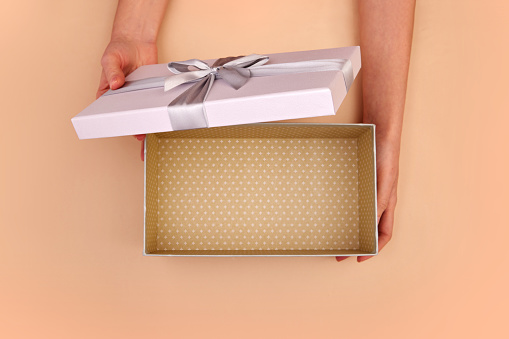 First person top view photo of hands unpacking craft paper gift box with gray satin ribbon bow view of the empty interior of a box on isolated pastel beige peach fuzz background with blank space