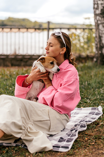 A young positive woman in a pink jacket and sunglasses hugging friendly Jack Russell Terrier dog, enjoying happy moments together while walking in the park on a summer day.