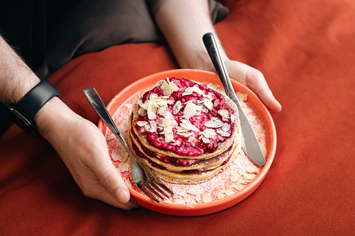 Appetizing pancakes with raspberry jam and almond flakes on a plate in male hands in bed, morning concept.