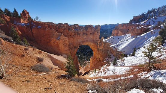 This stunning photo captures a natural rock arch in Bryce Canyon National Park, Utah. The red rock formation stands out against the snow-covered landscape and blue sky. The sunlight creates a beautiful contrast, highlighting the arch and the surrounding cliffs. This is a must-see destination for nature lovers and adventurers.