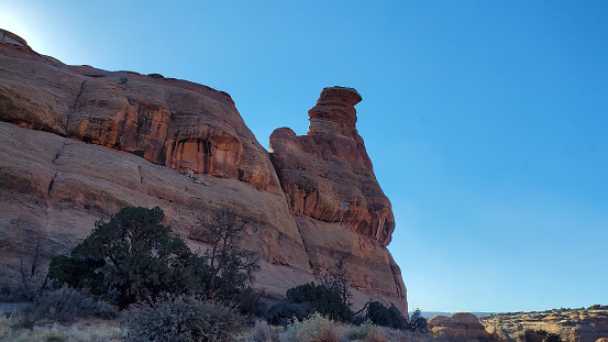 This photo showcases the stunning Gooney Bird rock formation in Moab, Utah. The red sandstone spire rises from the desert landscape. A sentinel for the off-road community who believe it is “good luck” to drive over the Gooney Birds toes before heading in to the wilderness. The clear blue-sky contrasts with the rugged terrain, creating a scenic and adventurous view. This is a unique and natural landmark in the American West, perfect for hiking, travel, and tourism.