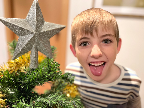 Siblings decorating the Christmas tree making funny faces. Homemade space. Children aged 7 and 10.  Funny moments with funny faces next to the Christmas star.  Real moments. Family. Warm tones.