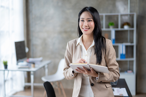 Beautiful Asian businesswoman secretary working happily in office using laptop and diligently taking notes.