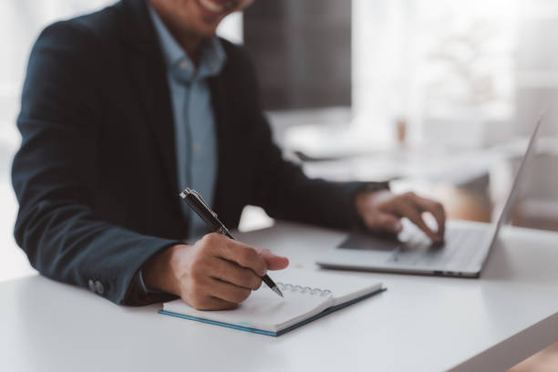 istock Closeup shot of businessman hand using pen and writing in notebook, presentation, working project at workplace. Man taking notes, typing on keyboard. 1847834414