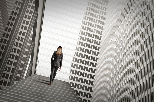 A businesswoman  climbs a set of concrete steps.  The steps are surrounded by the high rise buildings of a madern city.