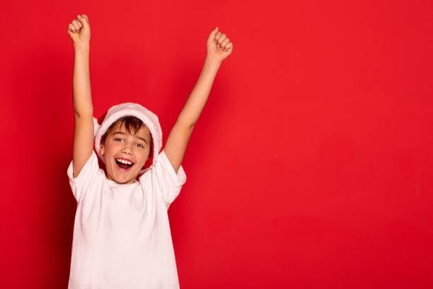 Happy smiling boy posing in Santa Claus hat on red studio background.. Lucky kid celebrating Christmas time.