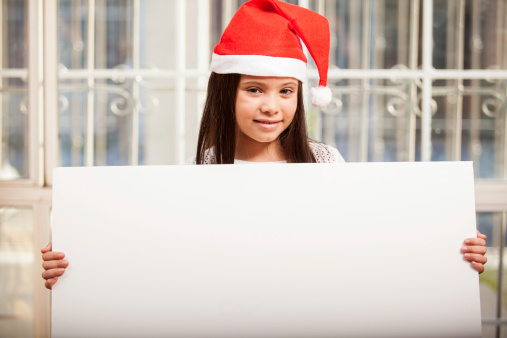 Happy little girl wearing Santa's hat and holding a big sign at home