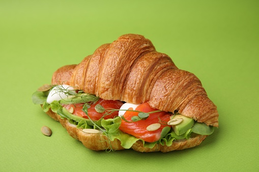 Tasty croissant with salmon, avocado, mozzarella and lettuce on green background, closeup