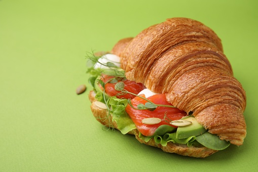 Tasty croissant with salmon, avocado, mozzarella and lettuce on green background, closeup. Space for text