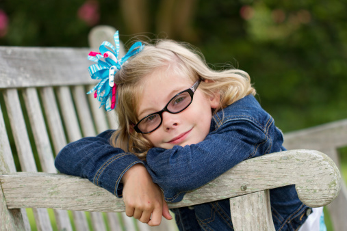 Young girl with a bow in her hair on a rustic park bench daydreaming. She is wearing a blue jean jacket and wears glasses.