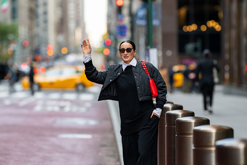 Well-dressed businesswoman wearing all black with white shirt, red bag and sunglasses seen standing on the street of Manhattan, New York and stopping a taxi while heading to work during one autumn day.