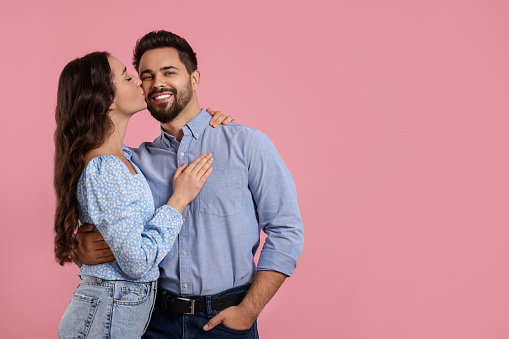 Woman kissing her smiling boyfriend on pink background. Space for text