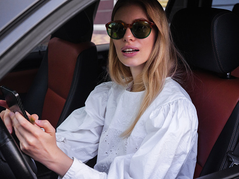 A young blonde girl enjoys talking with a friend on the phone while sitting in a car.