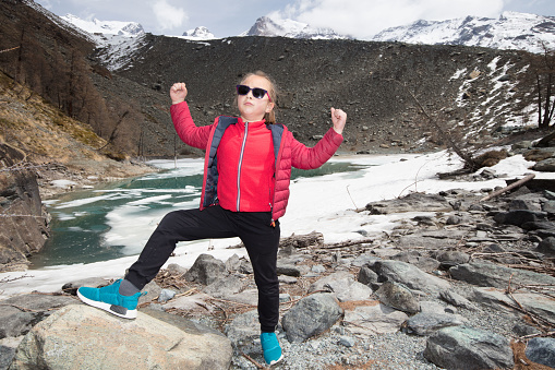 The mountain lake at the foot of the Monte Rosa massif, surrounded by steep boulders and glacier moraine, melts in spring. A young girl rejoices triumphantly at her achievement after a long hike
