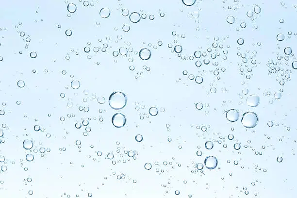 An underwater photograph of bubbles on a clear blue background.  The bubbles are randomly dispersed.  The bubbles vary in size with most being very small and a few being larger.  The background is a translucent blue.  The light comes from the bottom of the photo, making the bottom of the background very light blue, while the top is a darker shade.  The water is free of any visible impurities.  The bubbles seem to be largely concentrated in the upper right portion of the center of the photo, as well as the upper middle.