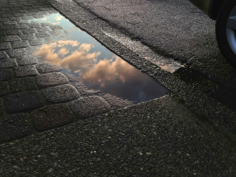 Reflection of clouds lit by setting sun in puddle on pavement by dark, wet curb and rear wheel of car. Camera: iPhone 4S.