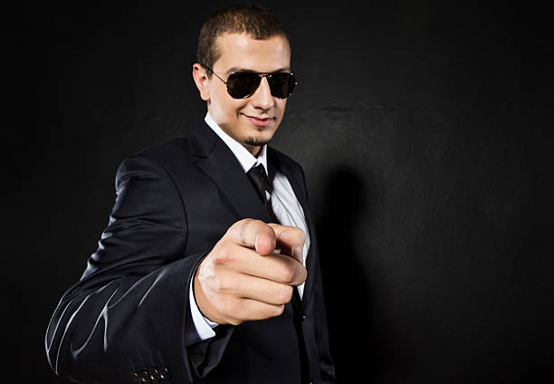 Young handsome man in black suit and glasses smiling and pointing isolated on black background