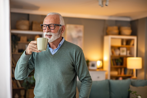 An elderly man is standing in the living room of a nursing home with a happy smile and holding a cup of coffee