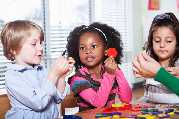 Multi-ethnic preschool children sitting at table in classroom playing with colorful wooden shapes (4 years).  Focus on girls.
