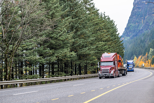 Convoy of the industrial standard big rig semi trucks with loaded semi trailers led by burgundy semi truck with flat bed semi trailer driving on the highway road with mountain and forest on the sides