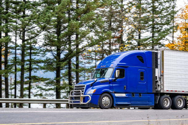 istock Streamlined shape blue big rig semi truck with grille guard transporting cargo in refrigerator semi trailer driving on the road along the river with autumn trees line 1848385546