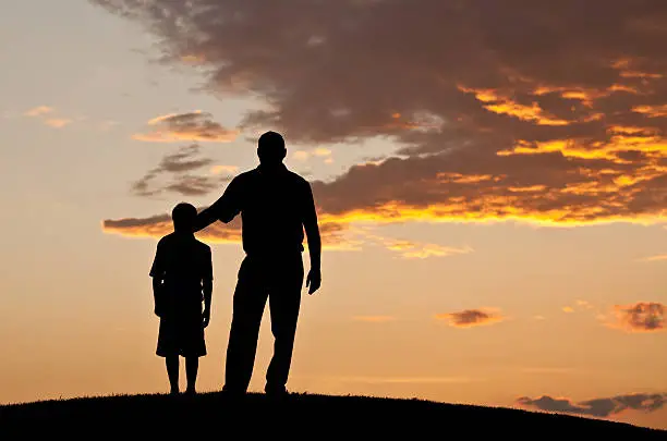 A father putting his arm around his son at sunset.  Standing on a hill. Silhouette. Additional themes include parenting, bonding, love, relationships, family, fathers, children, care, togetherness, support, love, single parenting, talking, consoling, and father's day. 