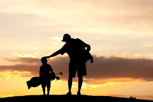 A junior golfer with his dad. Horizontal colour image. Silhouette. Golf concept. Additional themes in the image are sports, leisure, family, fun, mentor, coach, coaching, teaching, learning, junior level, son, boys, men, care, father, father's day, parenting, parent, single father, encourage, encouragement, love, relationships, and support. Boy is young and kindergarten age. 